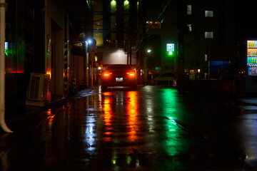 A moody nighttime photograph of a narrow wet street in Japan, illuminated by colorful artificial lights and their reflections on rain-soaked pavement. In the center, a dark-colored car with glowing © Andrius