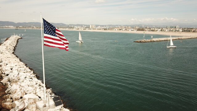 Aerial view of the American flag waving on a rocky breakwater with sailboats navigating the harbor entrance in Long Beach, California, United States.