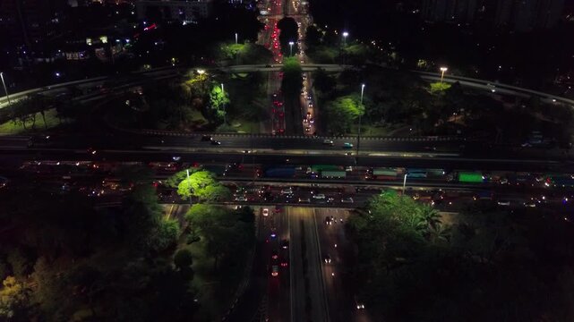 Aerial night view of a curved ramp section of the Semanggi Interchange in Jakarta, Indonesia, with city lights and vehicle headlights visible through the surrounding tree canopy.