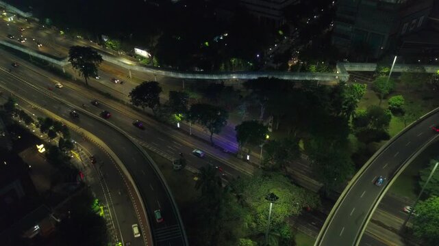 Aerial night view of a curved ramp section of the Semanggi Interchange in Jakarta, Indonesia, with city lights and vehicle headlights visible through the surrounding tree canopy.