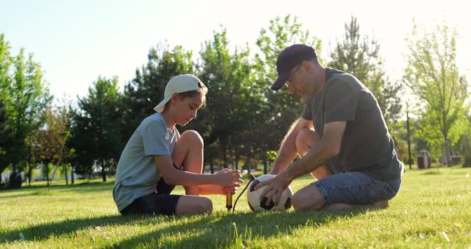 Father and son preparing soccer ball before game on green grass at sunset. Dad helps with equipment while boy pumps air into ball. Learning by example, practical skills and active parenting