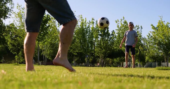 Barefoot father and son pass soccer ball on green grass in park at sunset. Close up on father's feet while child plays in background, capturing family bonding and outdoor activity.