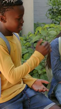 Vertical video: Leaning, boy in yellow shirt offering and gesturing on bench, light-gray backpack