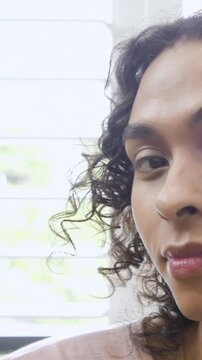 Vertical video: Looking cam nonbinary dabbing paint on cheek near blinds checking brush, copy space