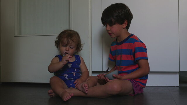 Siblings sitting on kitchen floor sharing food from bowl expressing playful interaction bonding and relaxed family moment during everyday indoor meal time activity