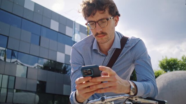 Man pauses with smartphone near bicycle in front of glass office