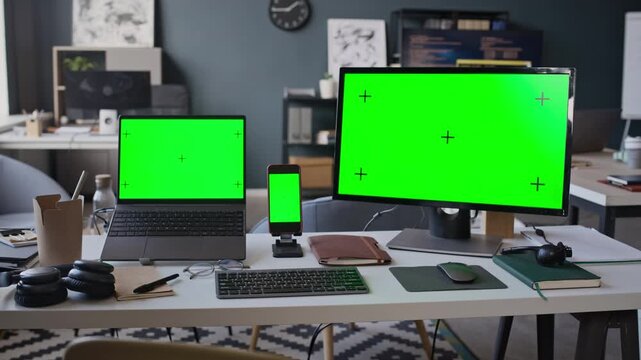 Zoom in shot of multiple devices including laptop, smartphone, and desktop monitor displaying green screens on office desk. Perfect for displaying software mockups