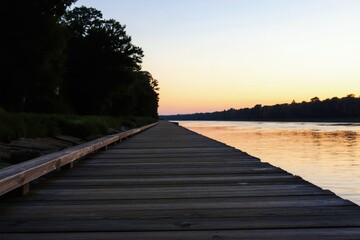 Obraz premium Wooden walkway along calm river at twilight. Trees line the shore under a soft sky. Peaceful path leads to horizon, reflecting orange light.