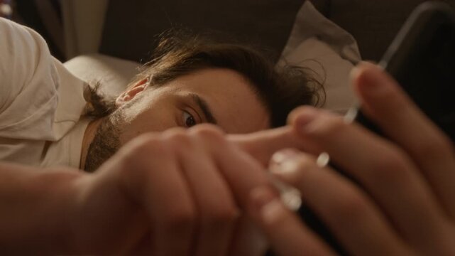 Young Caucasian man lying in bed and scrolling on smartphone screen after waking in morning. For commercials promoting mobile applications, sleep tracking technology, or social media platforms
