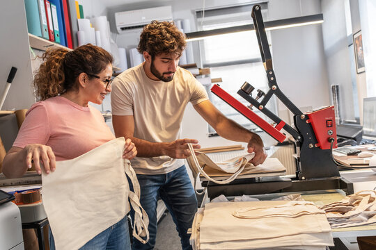 A print shop worker is operating a heat press machine for textile printing, carefully aligning the material before pressing. The scene shows a typical production step in custom garment printing with f