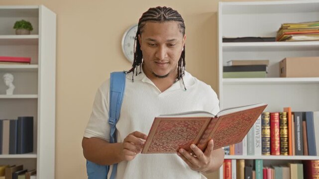 Man with hands holding an ornate book, backpack on shoulder, reading and turning pages in an indoor library room with bookshelves and wall clock; focused study.