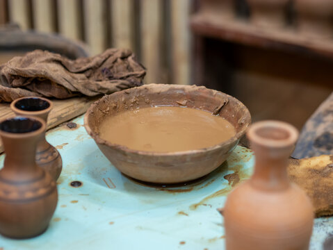 A rustic earthenware bowl filled with liquid clay slip sits on a messy workbench surrounded by terracotta vases in a pottery studio.