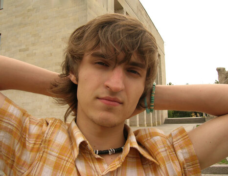 Street city photo. Young adult white man with checkered cotton orange shirt with short sleeves, blue bracelet on his hand, black coral beads on his neck looking at the camera. Concept of rest, trip