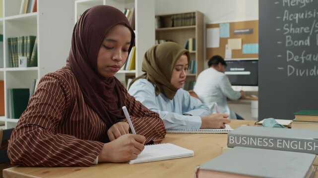 Young adult Asian female students wearing hijabs taking notes during Business English class in library. Clip is perfect for marketing campaigns on adult education, diversity, and language learning