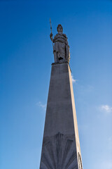 Obraz premium Statue of a female warrior with a spear on top of a tall stone monument against a clear blue sky