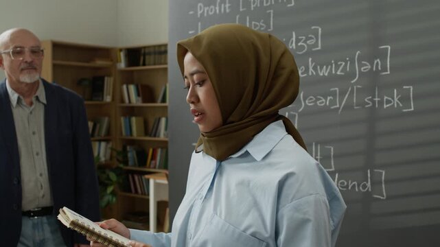 Asian young adult student wearing hijab holding notebook and speaking during presentation in classroom while senior Caucasian male teacher listens. Clip suitable for language school ads