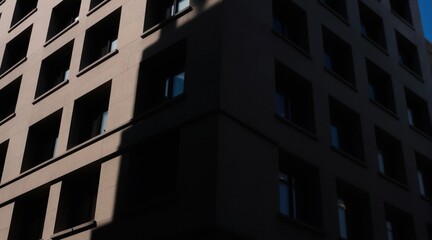 Obraz premium Building facade with geometric shadows and repeating windows. High contrast between light and dark areas creates a dramatic urban mood. Modern structure with minimalist design.