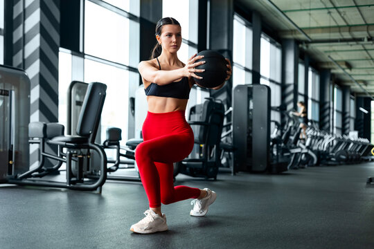 Athletic woman in red leggings doing forward lunge with medicine ball torso twist in gym