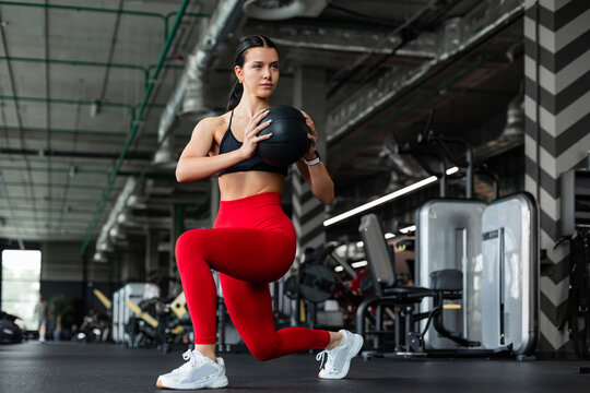 Athletic woman in red leggings doing forward lunge with medicine ball torso twist in gym