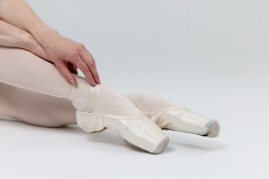 Ballerina sitting and tying satin ribbons on pointe shoes close up on white background