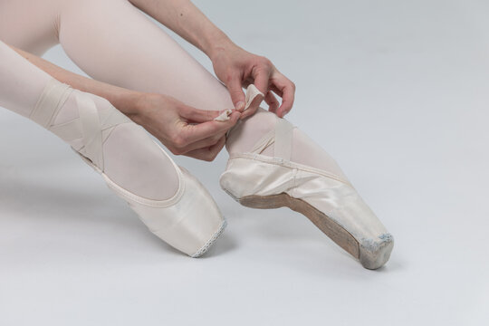 Ballerina sitting and tying satin ribbons on pointe shoes close up on white background