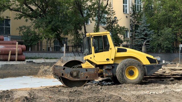 Heavy yellow soil compactor roller vehicle standing at an urban road construction site near a stack of industrial corrugated plastic pipes. Infrastructure development and street repair works.
