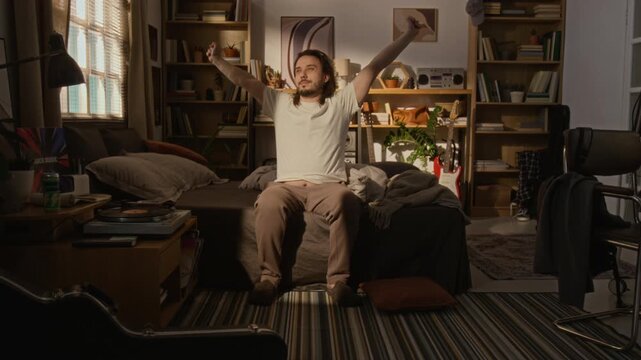 Freelance Caucasian young adult man sitting on bed in home studio, stretching arms and yawning after waking up. Footage is ideal for morning routine, remote work lifestyle, and wellness commercials