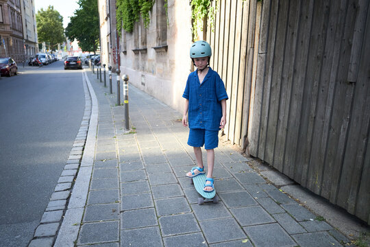 Young child in blue outfit and helmet rides a skateboard on a city sidewalk