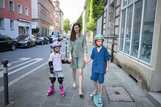 Mother walks with children on roller skates and skateboard down city street