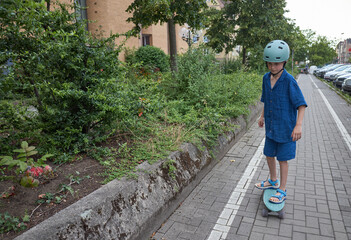 Young boy in helmet rides a skateboard on a paved path next to green bushes © 4Max