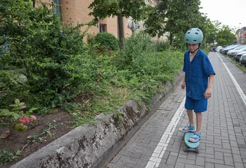 Young boy in helmet rides a skateboard on a paved path next to green bushes © 4Max