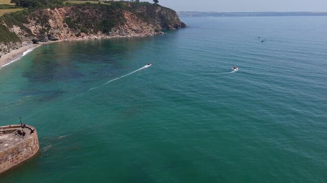 Aerial clip showing two small fast boats speeding across the vivid turquoise sea immediately after exiting the stone structure of Charlestown Harbour near a steep cliff line. Charlestown, United Kingd