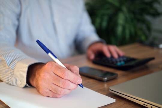 Man writes down financial notes and calculates expenses at desk using calculator, smartphone and laptop. Concept: budgeting, accounting and personal finance management.