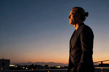 Fototapeta premium Man in dark suit stands on a balcony during twilight. Golden hour light illuminates his profile against an orange and blue gradient sky over distant city lights and mountains.