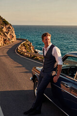 Fototapeta premium Man in formal waistcoat leans against a classic dark blue convertible car along a coastal road. Warm sunset light hits his face while the ocean waves crash nearby rocky cliffs.