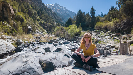 Fototapeta premium A young woman sits peacefully by a flowing river, surrounded by majestic mountains and lush greenery.