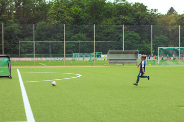 Obraz premium Young boy in sportswear runs towards a soccer ball on a green artificial turf field with goalposts and players visible in the background