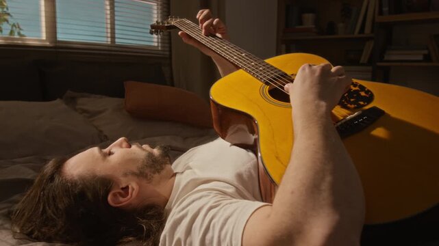 Young Caucasian male freelancer lying on bed and playing acoustic guitar for workday break. Commercial use in marketing campaigns promoting mental health, creative hobbies, or music education