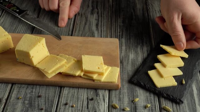 Hands slicing yellow cheese into thin squares on a wooden cutting board with a black slate plate and scattered spices in a kitchen setting
