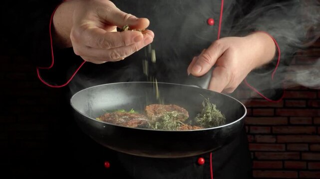 Chef in black uniform sprinkles dry herbs over sizzling meat in a frying pan, with steam rising and a cozy kitchen featuring a brick wall visible in the background