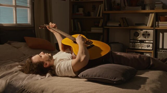 Young Caucasian man lying on bed in home studio, reaching for acoustic guitar and tuning strings. Footage is ideal for music education, creative lifestyle, or leisure marketing campaigns