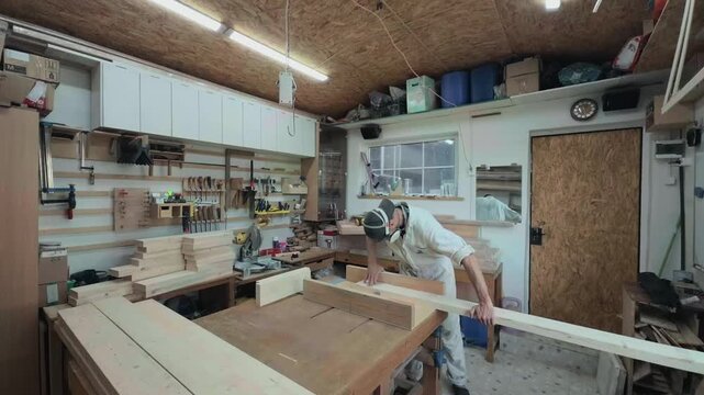 Authentic male joiner in protective respirator feeding a long timber plank into a table saw sled inside a rustic garage workshop wide angle tilt up shot of physical woodworking preparation
