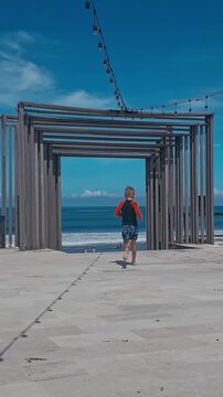 Boy Running Toward Sea Gate On Sunny Coastal Promenade, Timber Arch Framing Ocean Horizon Under Clear Blue Sky, Back View Of Kid In Red Rashguard And Patterned Swim Shorts, String Lights Overhead,Bali