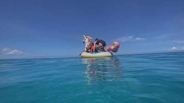 Two caucasian boys on inflatable unicorn pool float, mother guiding and laughing, bright blue water under clear sky, rainbow mane and colorful tail, playful splashes, sunlit family