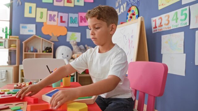 Boy places yellow wooden puzzle piece with right hand on red table amid toys and alphabet posters in a classroom building; concentration.