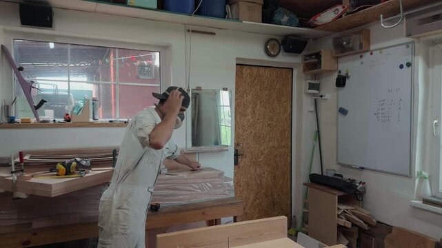Exhausted local laborer in dusty white coveralls taking off respirator mask and wiping sweat from face in bright rustic garage studio candid close up documentary shot