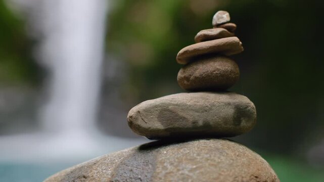 Carefully balanced stack of smooth river stones creates a peaceful zen cairn against a soft focus waterfall and lush green forest backdrop. Tranquil meditation scene in natural setting.