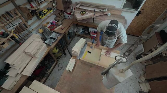 Unrecognizable cabinetmaker in safety respirator securing lumber strip with a blue clamp and dividing timber on a table saw inside a cluttered maker space overhead bird's-eye perspective shot