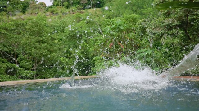 Slow motion footage of a young boy leaping into a turquoise infinity pool surrounded by lush tropical jungle. Water splashes dramatically as he lands and swims in the crystal clear water.