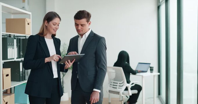 Portrait of young businesswoman shows digital tablet to man colleague in office. Woman manager discusses project realization with partner in corporate workplace